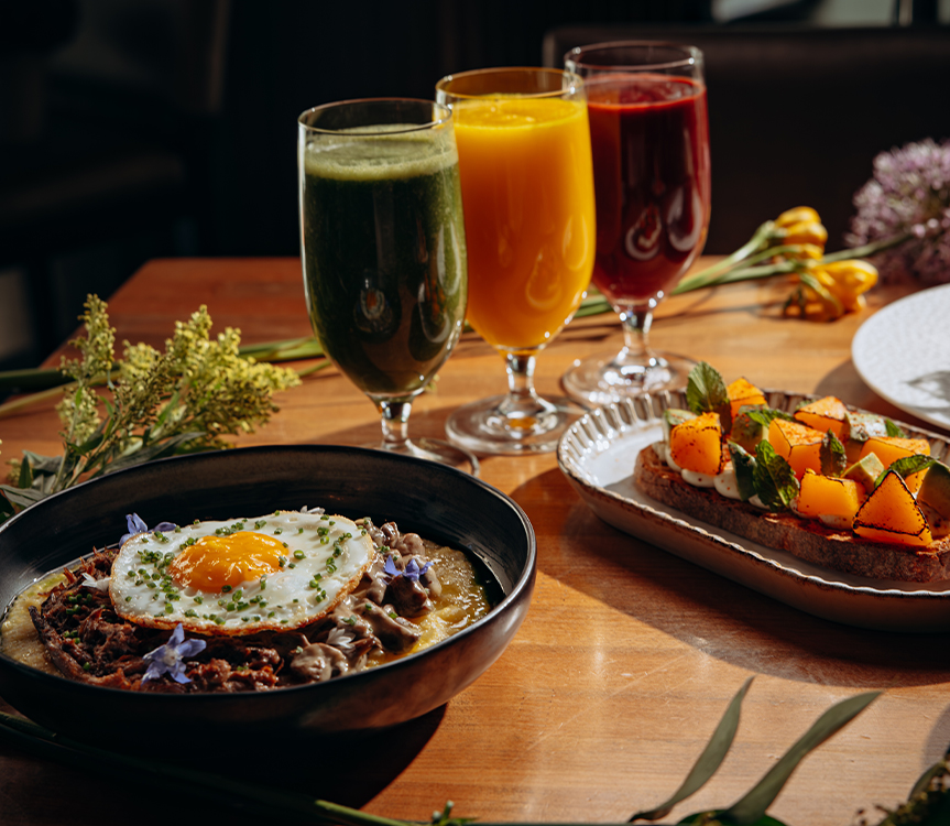A table set with a variety of dishes and drinks, including a bowl of food with an egg on top, a plate of toast with vegetables, and three glasses of juice.