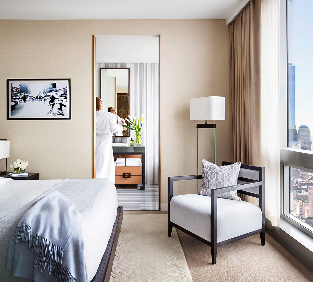 A hotel room with a bed, chair, lamp, and window showing a city view, with a person in a robe reflected in the mirror.