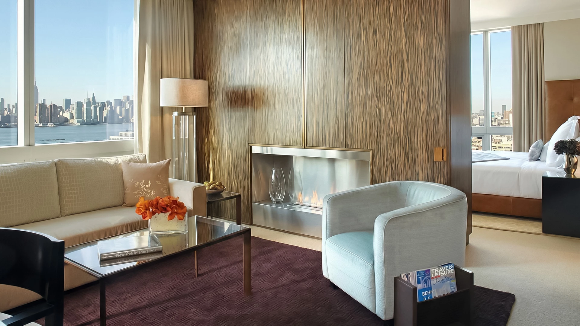 A luxurious hotel room with a city view, featuring a sofa, armchair, coffee table, and fireplace.
