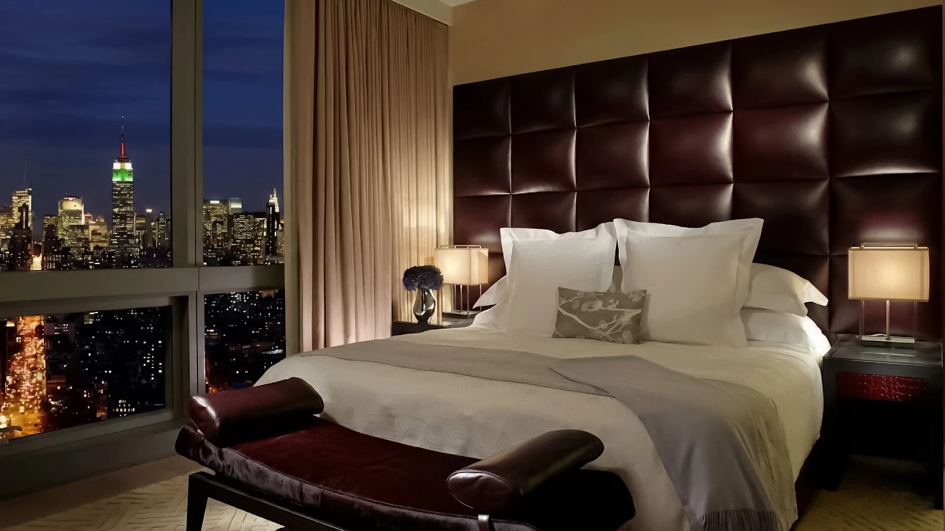 A hotel room with a large bed, leather headboard, and city view at night.