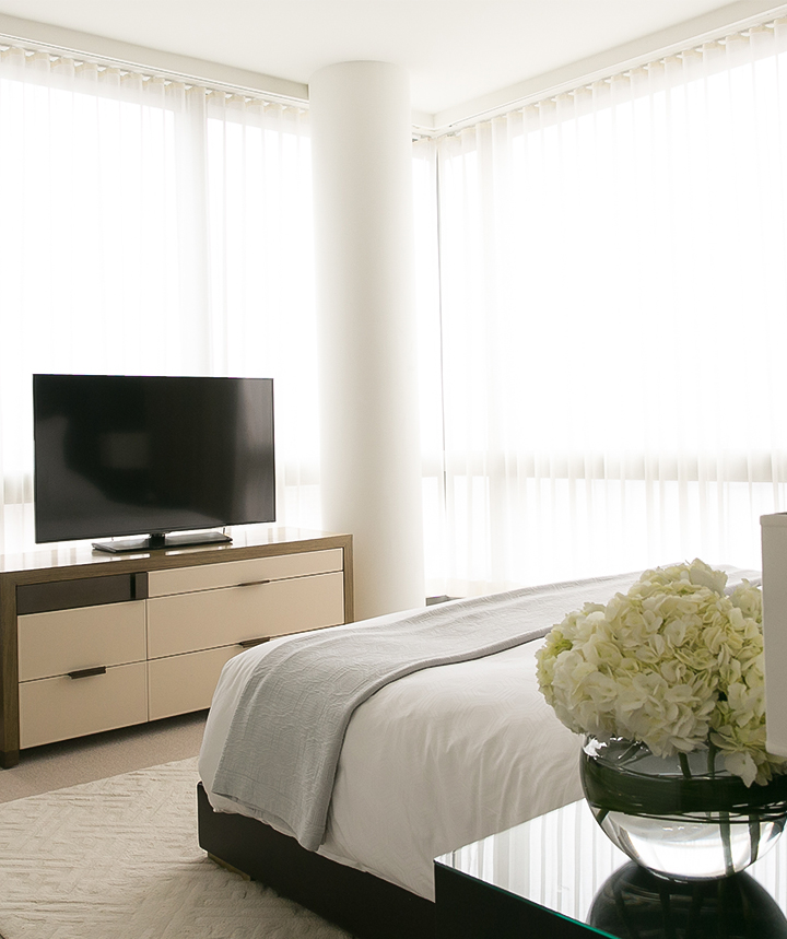 A hotel room with a bed, TV, dresser, and flowers on a nightstand in front of a large window.