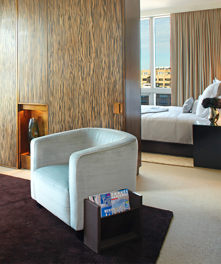 A hotel room with a light blue chair, a magazine rack, a bed, and a window.