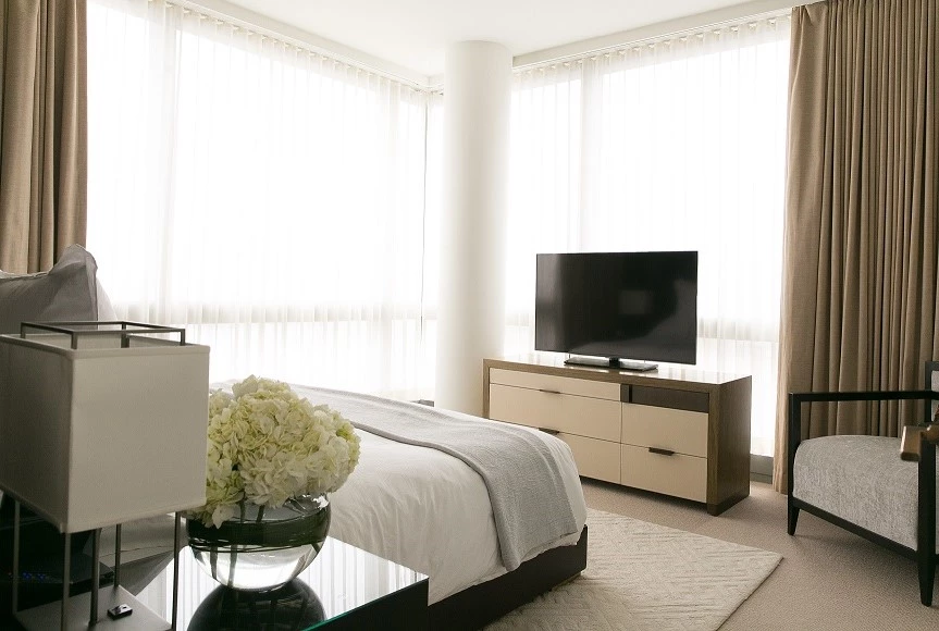 A bright and airy hotel room with a bed, TV, chair, lamp, and flowers.
