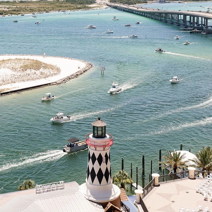 A lighthouse with a black and white diamond pattern stands in front of a body of water with boats and a bridge in the background.