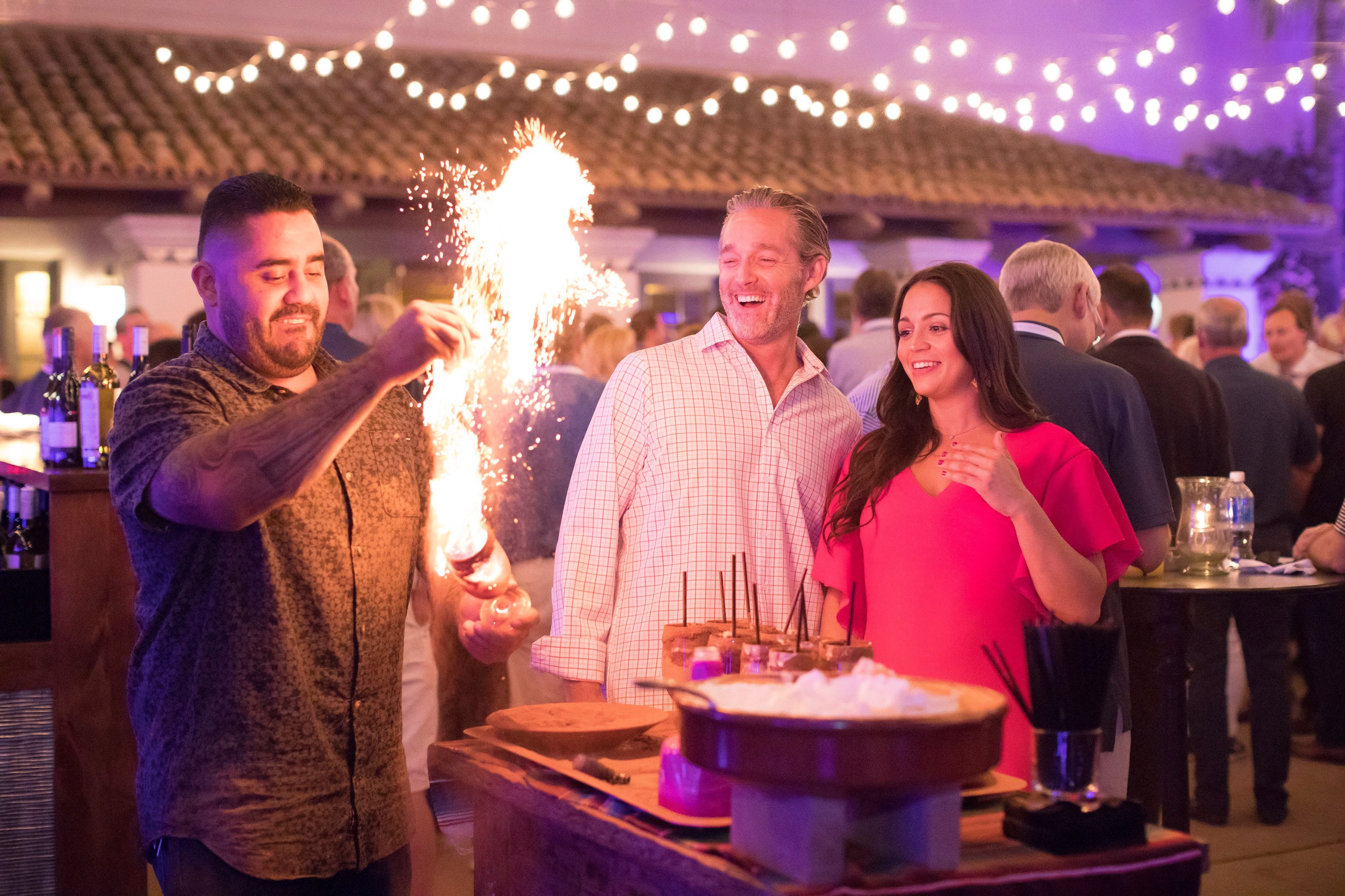 a person lighting a drink on fire and two guests looking and smiling