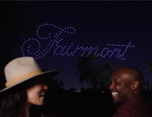 a man and woman smiling at night with drones behind them