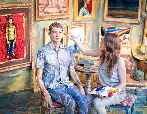 a smiling woman applying paint to a person with lots of artwork all around