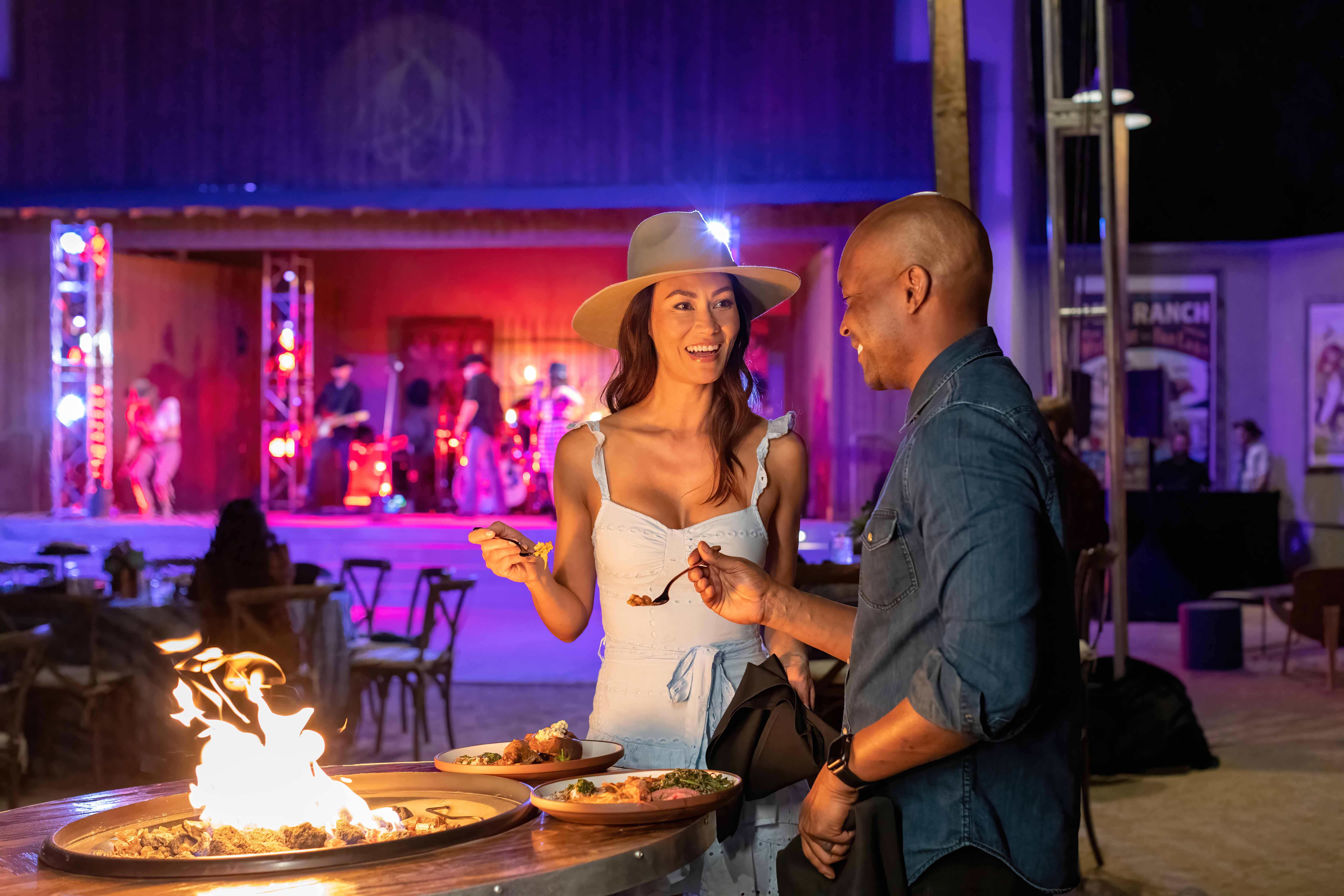 a man and woman standing next to a fire pit with food on it
