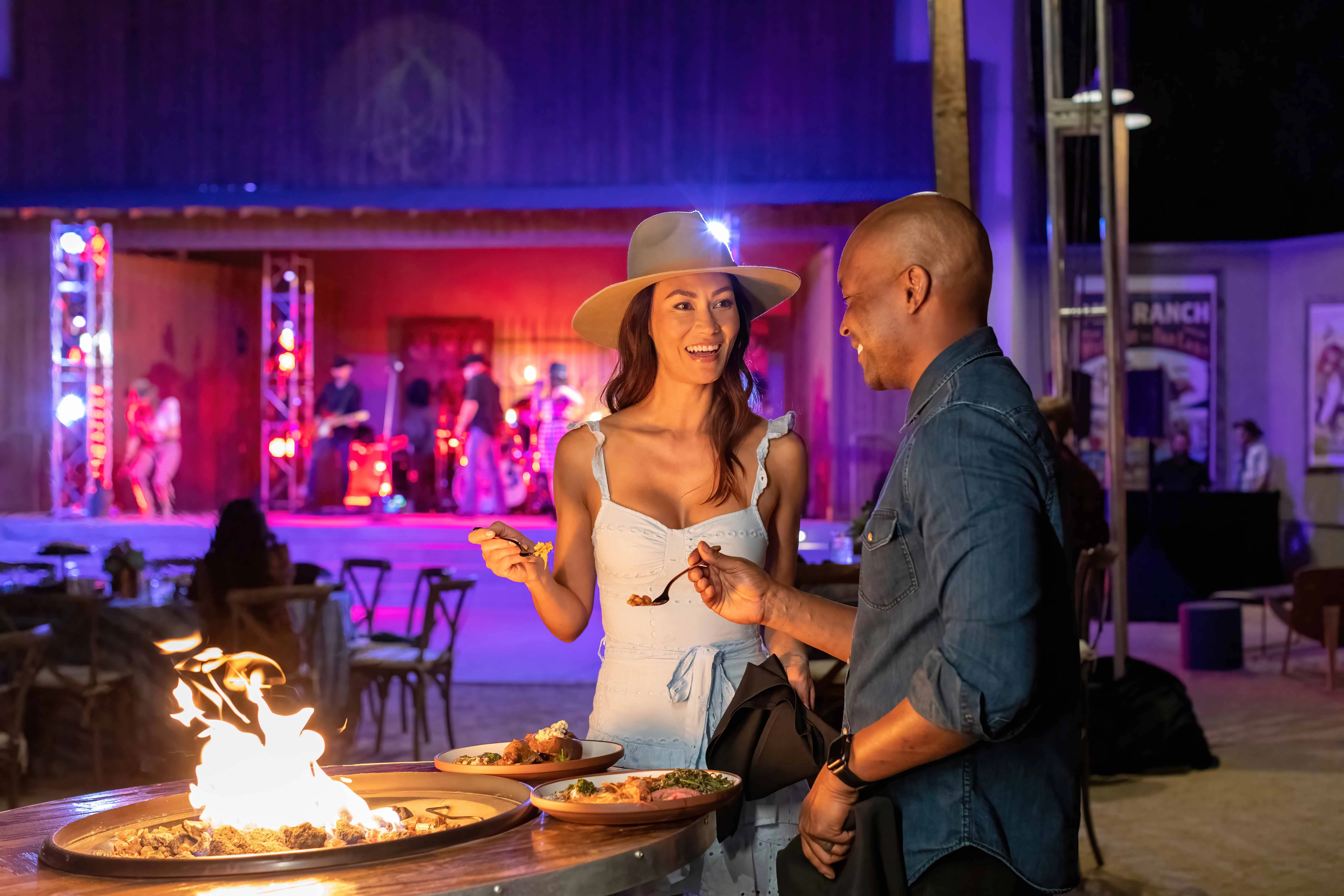 a man and woman standing next to a fire pit with food on it