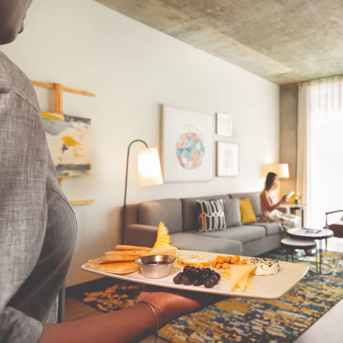 woman holding a plate of food walking towards a woman sitting on the couch