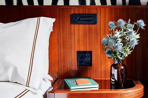 A close-up of a bed with a wooden headboard and nightstand, featuring white bedding, a book, and a vase with flowers.