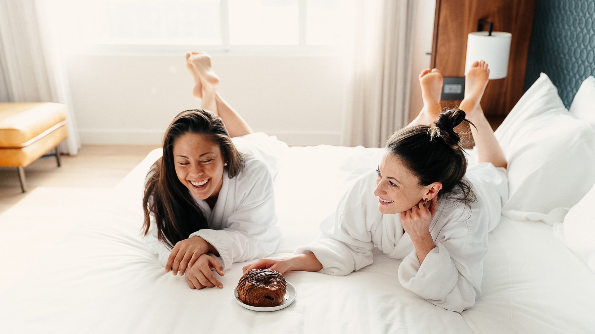 two women in white robes lying on a bed with a plate of food