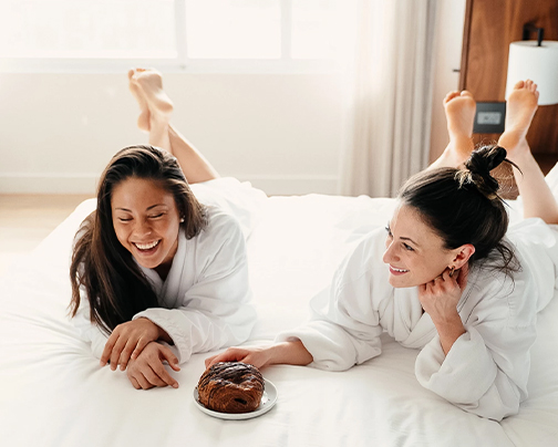 two women in white robes lying on a bed with a plate of food