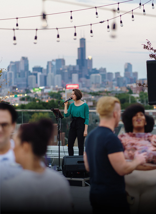 a woman singing into a microphone on a rooftop with a city in the background