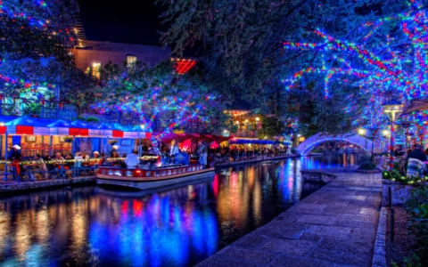 a river with a boat and lights with San Antonio River Walk in the background