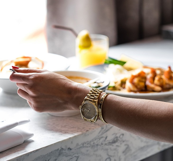A woman wearing a gold watch and bracelet sitting at a marble table eating food.