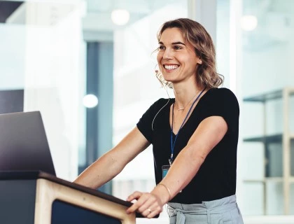 A smiling woman with light brown hair standing at a podium with an opened laptop.