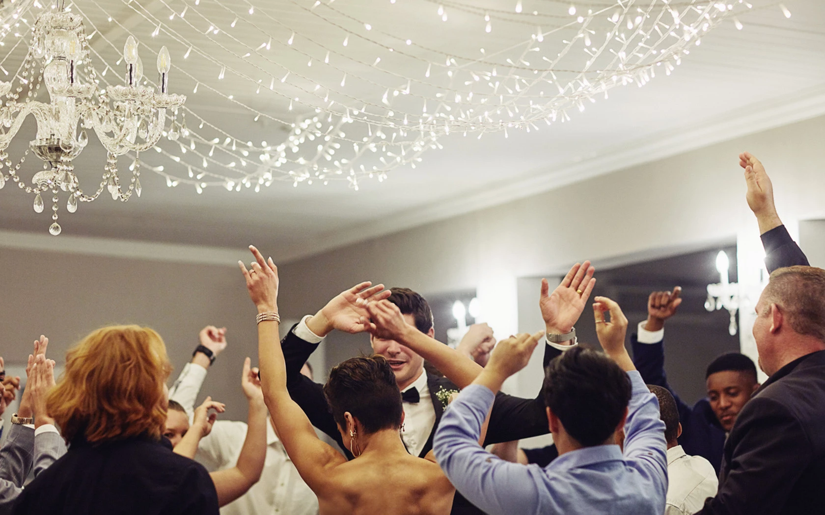 Wedding guests dancing with their hands held in the air and small string lighting hanging from the ceiling.