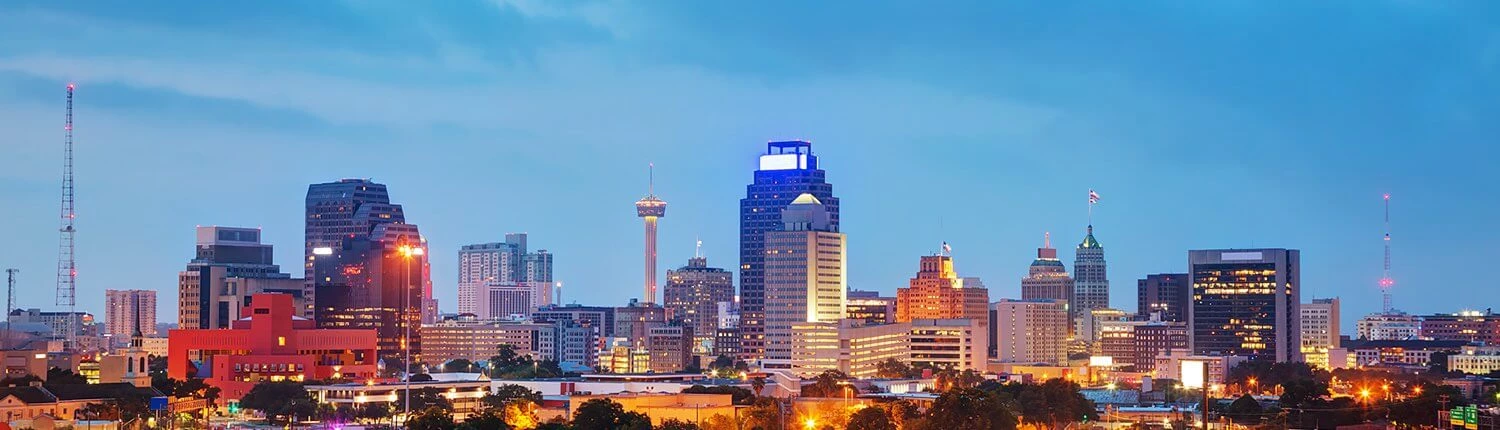 A city skyline at dusk with lit skyscrapers and a prominent tower in the distance.