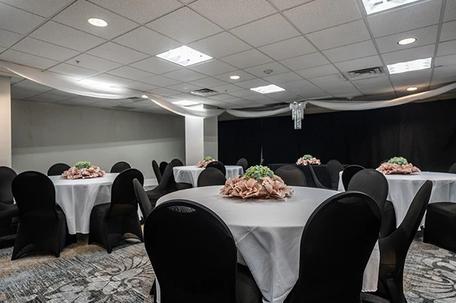 A meeting room with round tables set for an event, featuring white tablecloths, black chairs, and floral centerpieces.
