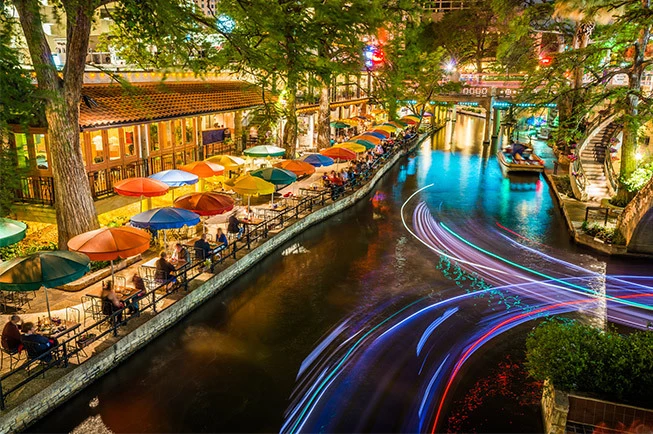 A nighttime view of the San Antonio Riverwalk with restaurants and people.