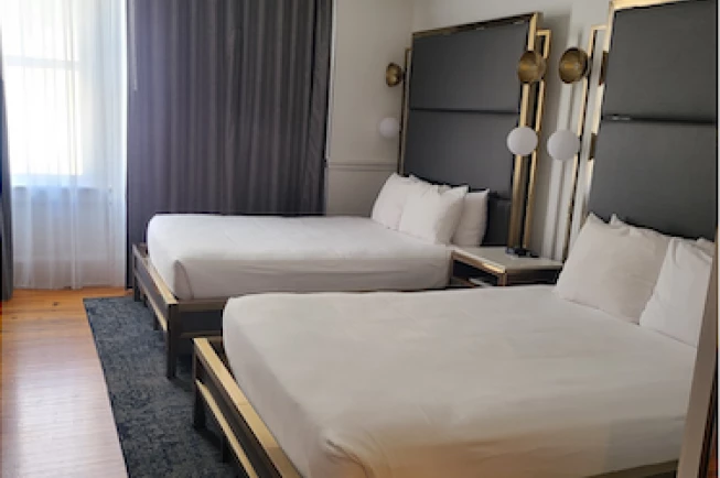 A hotel room with two twin beds, dark headboards with gold accents, and a window with sheer curtains.