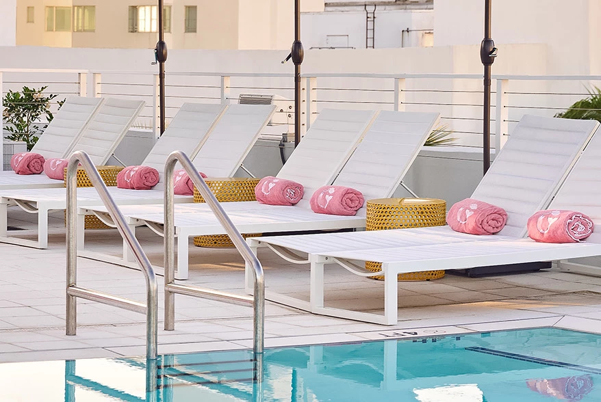 A row of white lounge chairs with pink towels beside a pool.