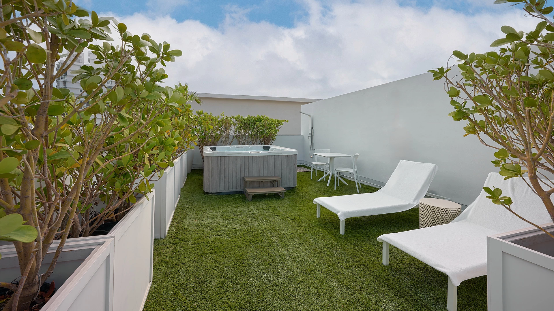 A small outdoor space with artificial turf, a hot tub, two lounge chairs, and trees in planter boxes.