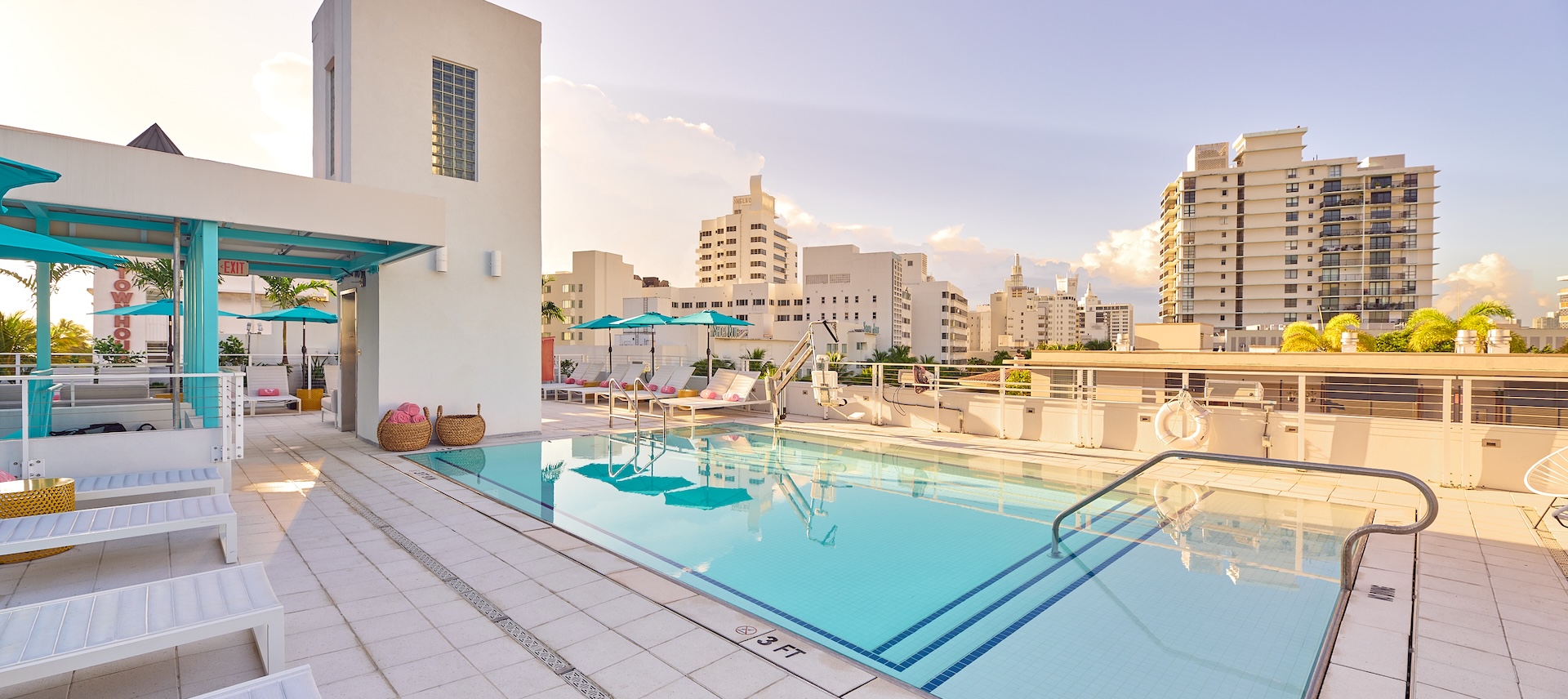 A rooftop pool area with a rectangular pool, lounge chairs, and umbrellas, overlooking the city skyline.
