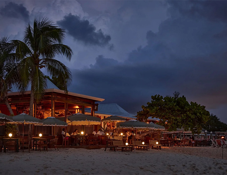 a beach with tables and umbrellas and a building with trees and a cloudy sky