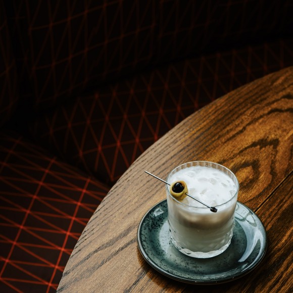 a glass of liquid with a olive on a round wooden table