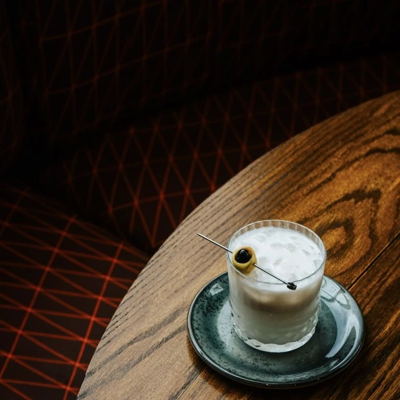 a glass of liquid with a olive on a round wooden table