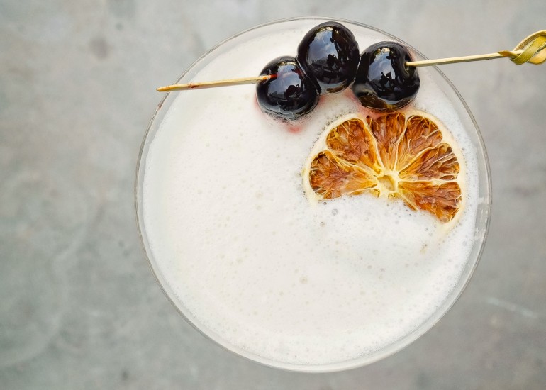 a glass of white liquid with a slice of orange and black grapes on top