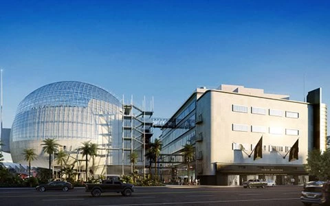 A beige hotel building with a glass dome structure to the left, palm trees in front, and cars driving by on a sunny day.