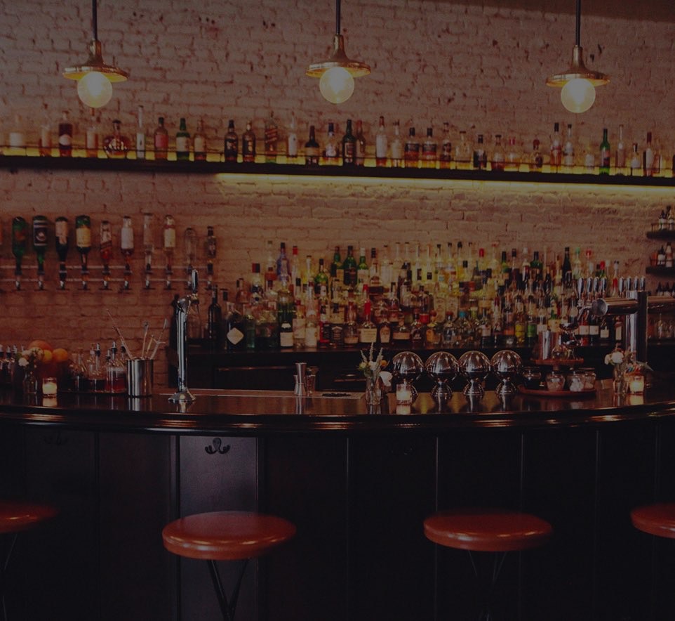 A dimly lit bar with a long counter, stools, and many bottles of liquor on shelves.