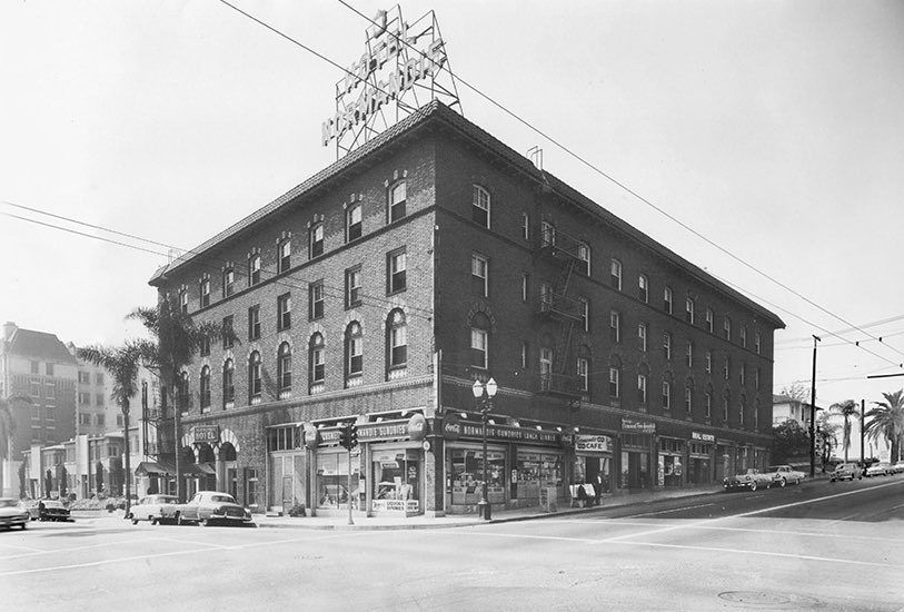 A black and white photo of a multi-story brick building on a street corner with shops and cars.