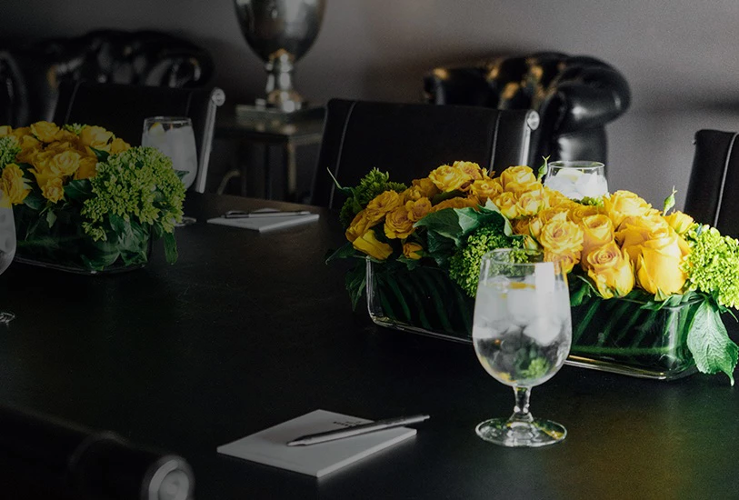 A meeting room table set with yellow roses, glasses of water, and notepads with pens.