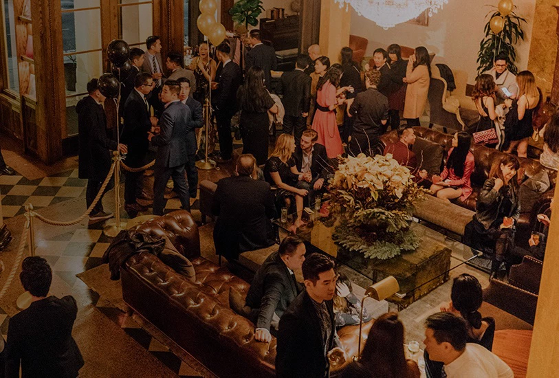 A large crowd of people gathered in a lobby with brown leather furniture, a checkered floor, and a chandelier.