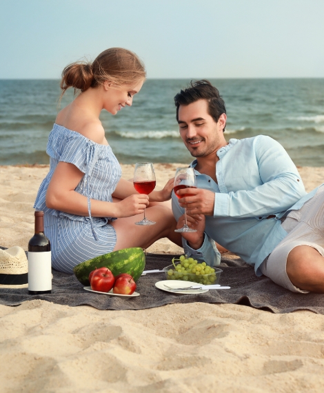 a man and woman sitting on a blanket on a beach with wine and fruit
