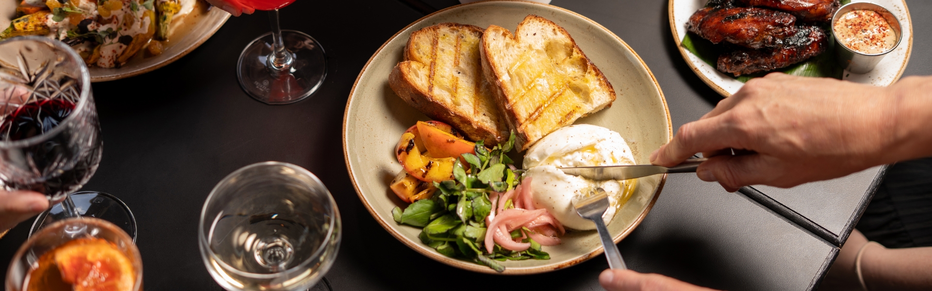 black table top with several dishes on top and person cutting into burrata