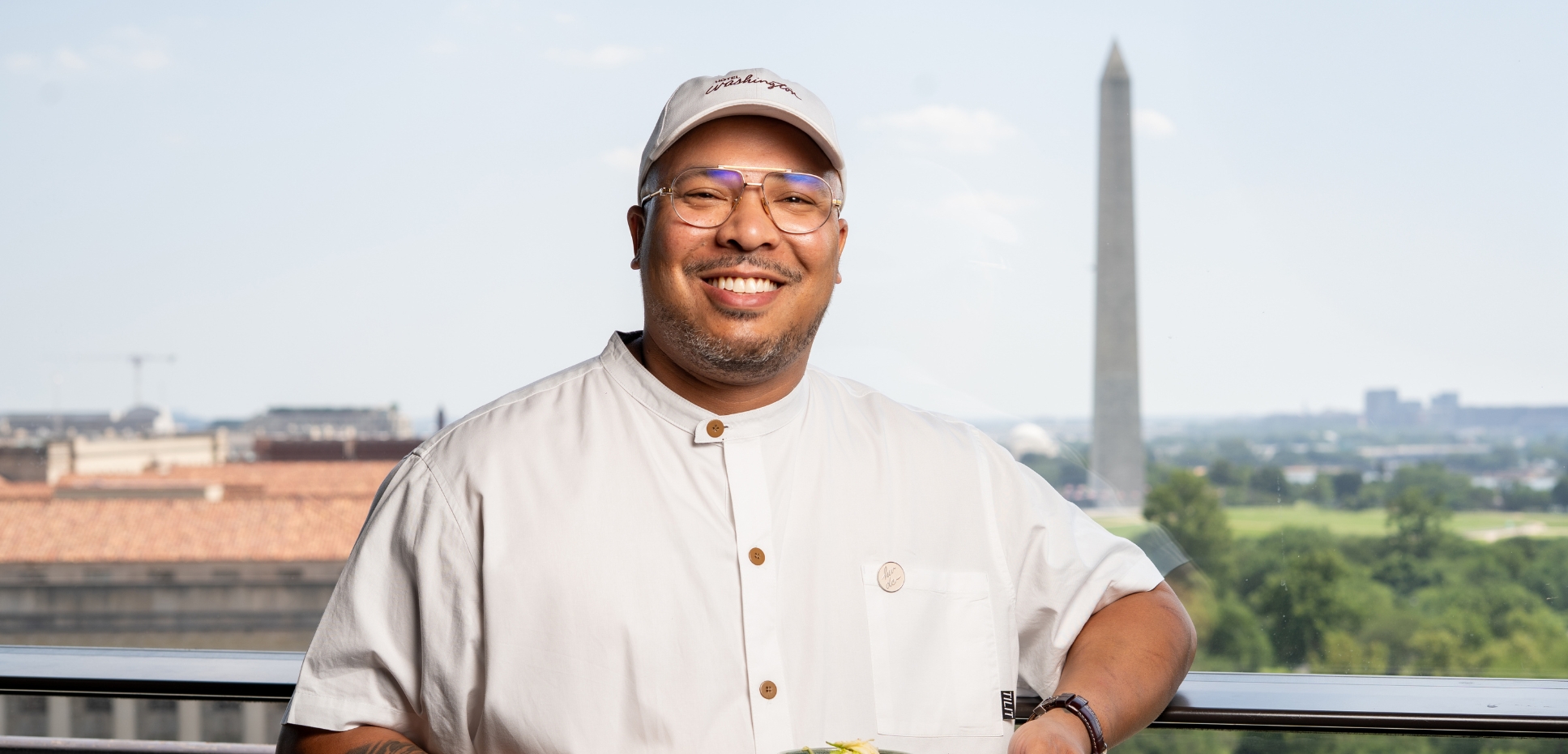 a chef wearing white jacket and baseball cap smiling and posing in front of window with the washington monument in background