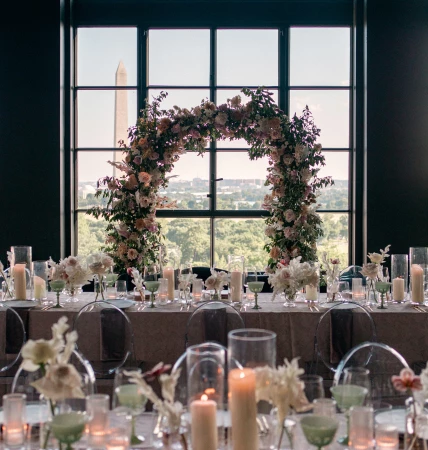 two tables set with pink table cloths and a floral arch in front of a large window