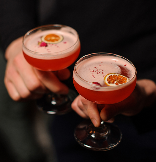 a pair of hands holding glasses with pink liquid and orange slices