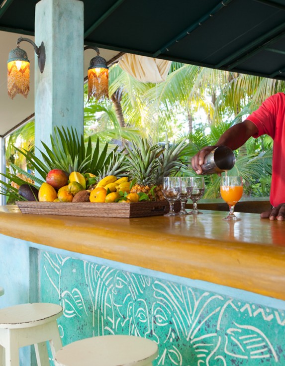A smiling bartender wearing a bright red shirt pouring a drink from behind the bar.