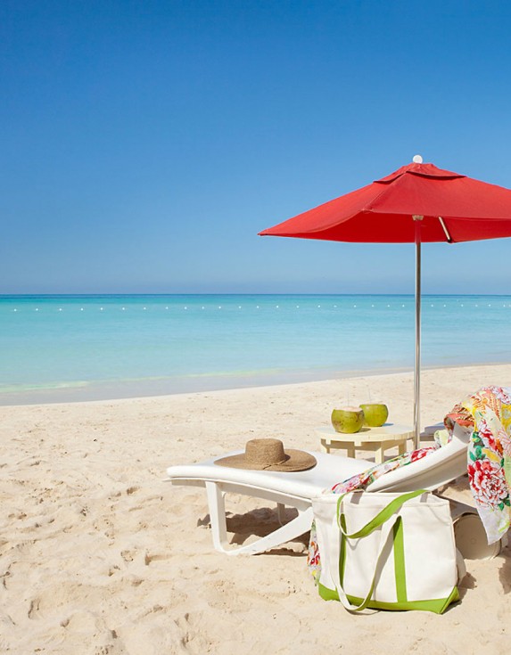 Crystal clear water and white sand with two lounge chairs under an opened red umbrella with the sun shining brightly.