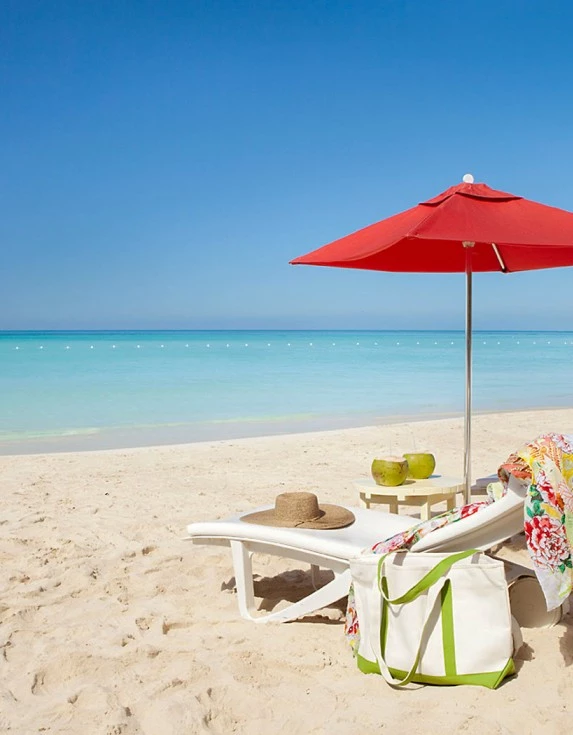 Crystal clear water and white sand with two lounge chairs under an opened red umbrella with the sun shining brightly.