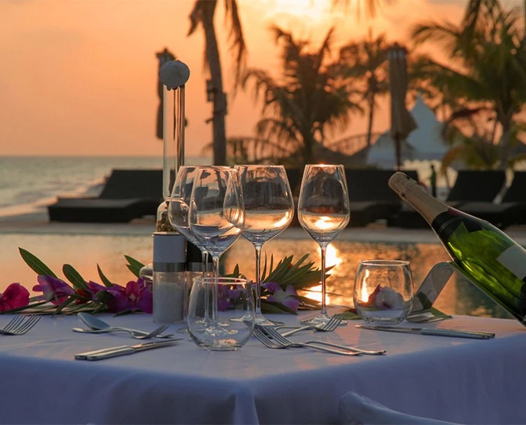 A white linen table set up for dinner service next to the water at sunset with palm trees in the distance.