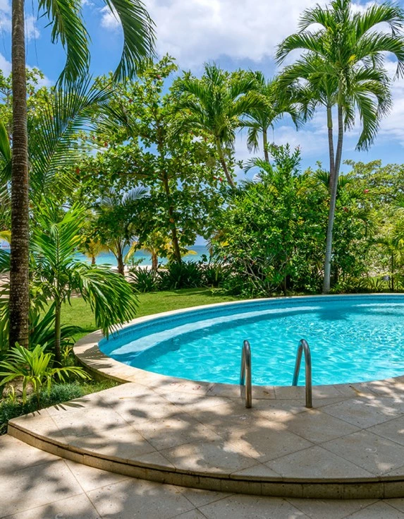 A blue pool next to the beach with a lot of tropical trees surrounding it.