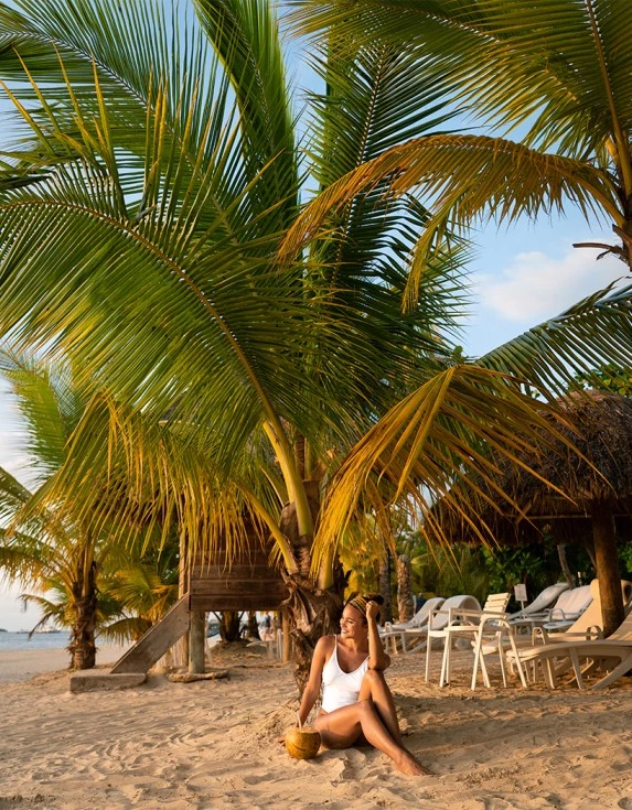 A woman wearing a white bathing suit sitting in the sand under a palm tree.