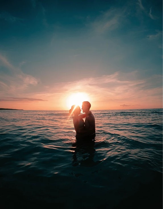 A man and woman hugging while standing in the water during the sunset.